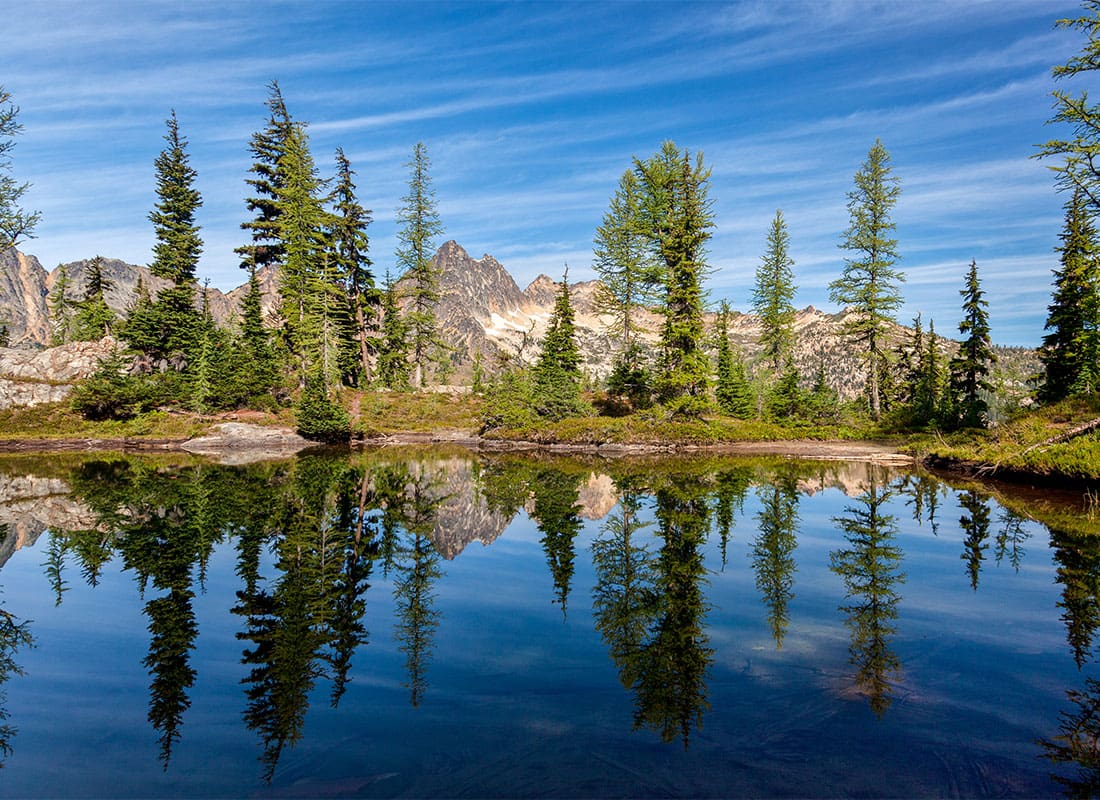Winthrop, WA - Pine Trees Next to a Lake with Reflections in the Water and Mountains in the Distance Against a Blue Sky in Winthrop Washington